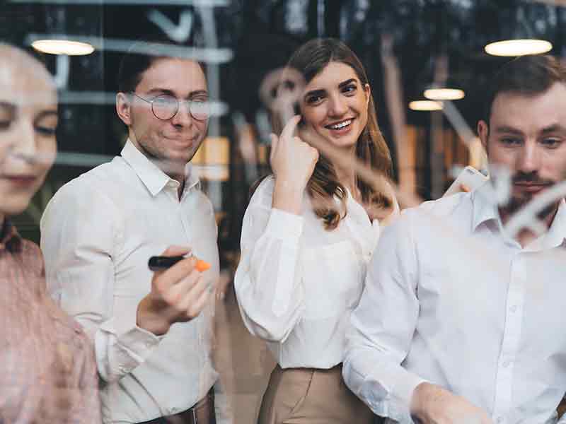office workers standing in front of a glass panel smiling and talking. One man has a whiteboard marker in his hand.